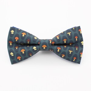 May include: A pre-tied bow tie featuring a dark teal background with a repeating pattern of small mushrooms in shades of orange, yellow, and brown. The bow tie has a structured, symmetrical shape with a central fabric knot and pointed ends.