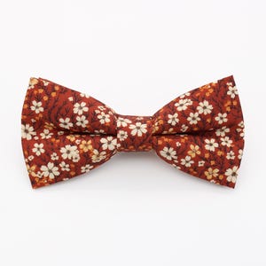 May include: A bow tie with a floral pattern. The bow tie is a deep reddish-brown color, with a repeating pattern of small white and yellow flowers. The bow tie is tied in a classic bow shape.