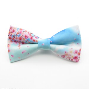 May include: A light blue bow tie with a pink cherry blossom print. The bow tie has a white background with pink flowers and light blue petals.