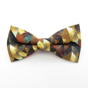 May include: A geometric patterned bow tie. The bow tie features a repeating pattern of gold, brown, teal, and red shapes. The bow tie is tied in the center and is set against a white background.