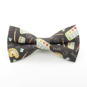 May include: A black bow tie with a pattern of paintbrushes, paint palettes, and the words "paints" and "paste" on it.