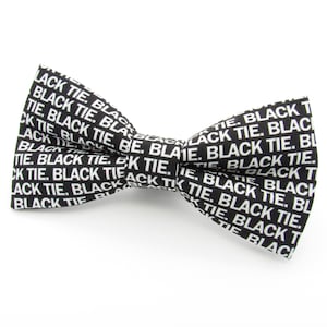 May include: A black bow tie featuring a repeating white text pattern that reads BLACK TIE. The bow tie has a classic shape with a central knot and two symmetrical wings, displaying the white sans-serif lettering clearly against the dark fabric.