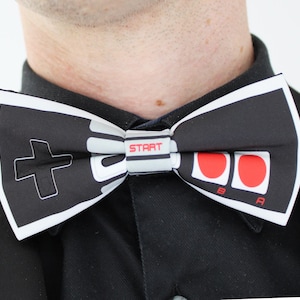 May include: A black and white bow tie featuring a graphic print of a retro video game controller. The design includes a black directional pad on the left, two red circular buttons labeled A and B on the right, and the word START in red text.