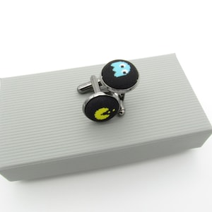 May include: A pair of round cufflinks featuring a black fabric surface with pixelated graphics. One cufflink displays a yellow circular character with an open mouth, and the other displays a light blue ghost character with white eyes.