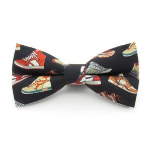 May include: Black bow tie with a pattern of colorful sneakers. The bow tie is made of fabric and has a classic bow tie shape.