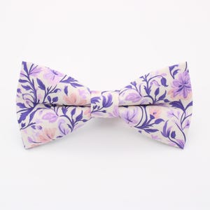 May include: A cream-coloured bow tie with a floral pattern in shades of purple and pink. The bow tie is knotted in the centre. This fashion accessory features a botanical design and is ready to wear.