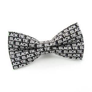 May include: A black bow tie featuring a repeating white text pattern that reads BLACK TIE. The bow tie has a classic shape with a central knot and two symmetrical wings, constructed from a fabric with a crisp, high-contrast graphic print.