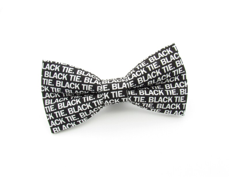 May include: A black bow tie featuring a repeating white text pattern that reads BLACK TIE. The bow tie has a classic shape with a central knot and two symmetrical wings, constructed from a fabric with a crisp, high-contrast graphic print.