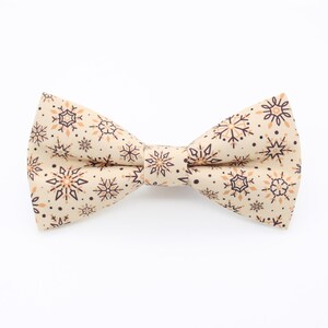 May include: A cream-coloured bow tie featuring a repeating pattern of snowflakes in shades of brown and tan. The bow tie is centred and appears to be made of fabric. The background is plain white.