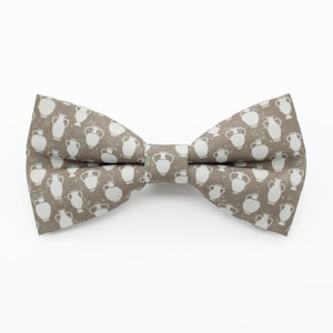 Può includere: Un papillon marrone con un motivo ripetuto di illustrazioni di vasi bianchi. Il papillon è realizzato in tessuto ed è legato a forma di fiocco classico. Lo sfondo è bianco.