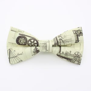 May include: A cream-coloured bow tie with a repeating pattern of vintage industrial machinery blueprints in grey and black. The bow tie is tied and ready to wear, suitable for a formal event or adding a unique touch to an outfit.
