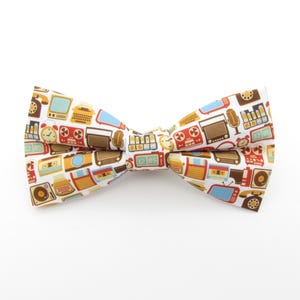 May include: A white bow tie with a repeating pattern of vintage electronics, including radios, televisions, and telephones in brown, blue, and orange. The bow tie is knotted in the center and is suitable for formal or casual wear.