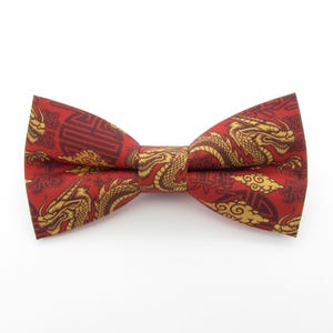 May include: A red bow tie featuring a repeating pattern of gold-coloured dragons, stylised clouds, and dark red geometric symbols. The fabric has a smooth texture and the bow tie is constructed with a central knot connecting two symmetrical wings.