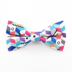 Può includere: Un papillon colorato con un motivo geometrico in tonalità di blu, rosa, verde e arancio. Il papillon è realizzato in tessuto bianco con un motivo geometrico.