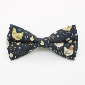 May include: A dark navy blue bow tie featuring a repeating pattern of yellow and white chickens, chicks hatching from eggs, and small yellow flowers. The fabric has a smooth texture and the bow tie is shaped with a central knot detail.