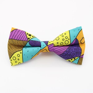 Può includere: Un papillon colorato con un design patchwork nei toni del giallo, viola, turchese e arancione. Il papillon ha una forma classica ed è realizzato in tessuto. Il design comprende varie forme e motivi, creando un accessorio unico.