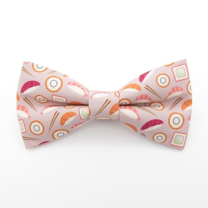 May include: A pink bow tie with a sushi pattern. The pattern includes sushi rolls, chopsticks, and shrimp.