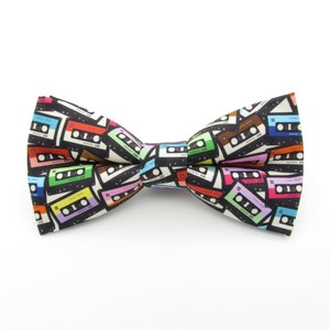 May include: A black bow tie featuring a pattern of colourful cassette tapes. The bow tie is made from fabric and has a satin finish.