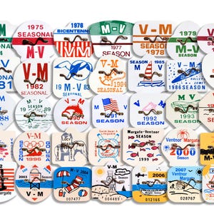 May include: A collection of 25 round, white plastic badges with various designs and text. The badges feature a variety of themes, including seashells, dolphins, sailboats, and American flags. Each badge has a metal hook on the top.