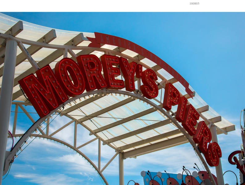 K&ouml;nnte beinhalten: Ein rotes Neon-Schild mit wei&szlig;en Lichtern, auf dem "Morey's Piers" steht, ist an einer wei&szlig;en Metallkonstruktion mit einem durchsichtigen Plastikdach angebracht. Das Schild ist gew&ouml;lbt und der Hintergrund ist ein strahlend blauer Himmel mit wei&szlig;en Wolken.