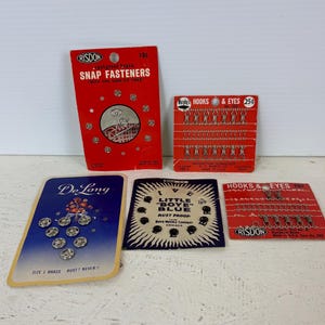 Puede incluir: Cuatro tarjetas de botones a presión, ganchos y ojales y botones vintage. Las tarjetas son rojas, azules y blancas con texto negro. Las tarjetas están etiquetadas como "Risdon", "De Long", "Little Boye Blue" y "Hooks & Eyes".