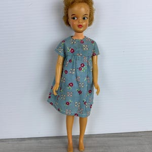 Könnte beinhalten: Eine Vintage-Puppe mit rotbraunem Haar und blauen Augen, bekleidet mit einem blauen Kleid mit Blumenmuster. Das Kleid hat kurze Ärmel und einen runden Ausschnitt. Die Puppe steht aufrecht und zeigt ihr klassisches Design.
