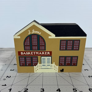 May include: A wooden house-shaped ornament with a black roof and mustard-yellow walls. The sign reads "BASKETMAKER" in red letters. The building has multiple windows with red trim and a small white porch. The name "Z. Jones" is above the sign.