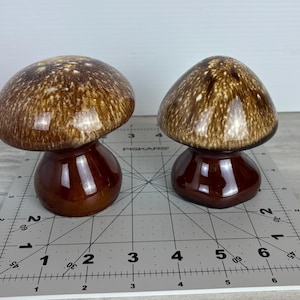 Puede incluir: Dos figuras de cerámica con forma de hongo. Los sombreros son marrones con motas más claras, y los tallos son de un marrón oscuro brillante. Las figuras se exhiben sobre una esterilla de corte gris con medidas.