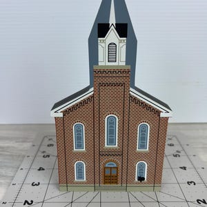 May include: A wooden church model with a red brick facade, arched windows, and a steeple. The model has a gray roof and a cross at the top of the steeple. The church has a brown door and is set on a gray surface.