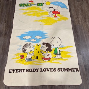 Puede incluir: Una toalla de playa beige con Snoopy y los personajes de Peanuts en la playa. La toalla presenta ilustraciones de Snoopy bajo una sombrilla y niños construyendo un castillo de arena. El texto incluye "COOL IT!" y "EVERYBODY LOVES SUMMER".