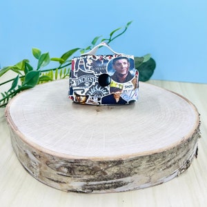 Puede incluir: Una pequeña caja blanca con un asa negra y un collage de imágenes y texto de la serie de televisión Supernatural. La caja está sobre una superficie de madera.