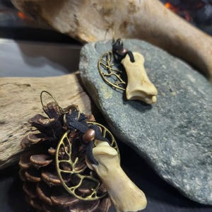 May include: A pair of unique earrings featuring bone pendants, accented with brass filigree and black cord. The earrings are displayed on a dark surface with a pinecone and a gray stone, creating a rustic aesthetic.