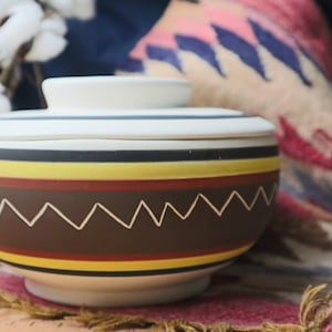May include: A white ceramic bowl with a lid, featuring a geometric pattern of brown, yellow, and red stripes. The bowl has a decorative design of white zigzags on the brown stripe.