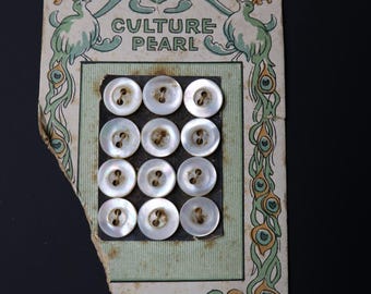 May include: Vintage card of Culture Pearl buttons. The card features a decorative border with birds and peacock feather designs. Twelve round, white buttons are arranged in a grid. The card has a torn edge.