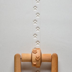 May include: A wooden rolling pin with a handle on each side. The rolling pin has a series of paw prints carved into the surface. The rolling pin is on a white surface with a line of paw print impressions.