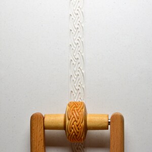 May include: A wooden rolling pin with a decorative, carved pattern. The rolling pin has a handle on each side and a removable roller. The roller is made of wood and has a carved pattern of a braided rope.