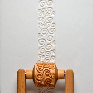 May include: A wooden rolling pin with a carved swirl design. The rolling pin has two wooden handles and is used to create decorative patterns in dough.