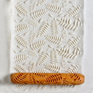 May include: A wooden rolling pin with a carved fern pattern. The rolling pin is on a white surface with a fern pattern embossed into it.