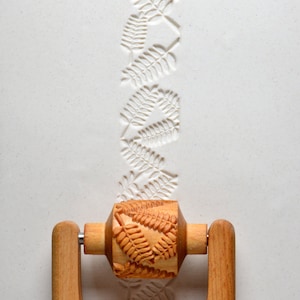 May include: A wooden rolling pin with a fern leaf design is used to create an embossed pattern on a white surface. The rolling pin has wooden handles and a carved design of fern leaves.