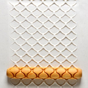 May include: A wooden rolling pin with a repeating pattern of rounded, overlapping shapes. The rolling pin is on a white surface with a raised, embossed pattern of a diamond-shaped net.