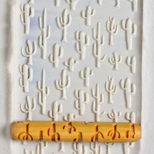 May include: A wooden rolling pin with a cactus pattern. The rolling pin is light brown and has a smooth surface. The cactus pattern is embossed on the rolling pin and is a light gray color.