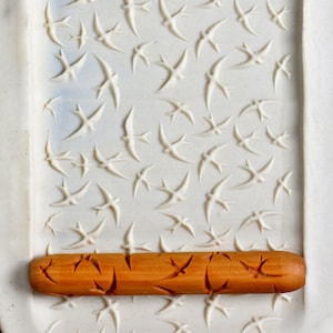May include: A wooden rolling pin with a carved design of birds and leaves. The rolling pin is on a white surface with a pattern of birds carved into it.