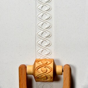 May include: A wooden rolling pin with a carved design of interlocking loops. The rolling pin has handles on either side and is resting on a white surface.