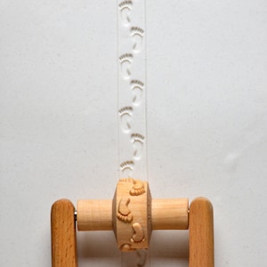 May include: A wooden rolling pin with a handle on each end. The rolling pin has a series of small, raised footprints carved into the surface. The rolling pin is used to create footprints in clay or dough.