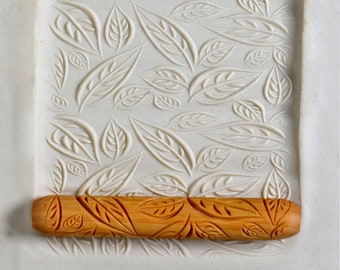 May include: A wooden rolling pin with a carved leaf design. The rolling pin is on a white surface with a leaf pattern embossed into it.