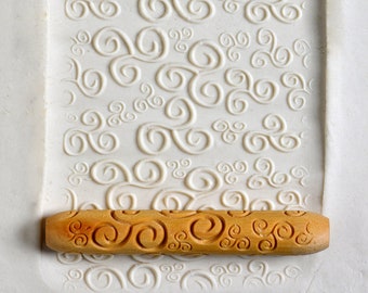 May include: A light-colored, textured rolling pin with a repeating swirl design, resting on a matching rectangular surface with an embossed swirl pattern. The rolling pin is made of wood and is approximately 10 inches long.
