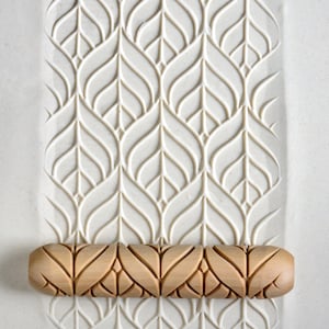 May include: A wooden rolling pin with a repeating pattern of leaves and swirls. The rolling pin is on a white surface with a similar pattern embossed into the surface.