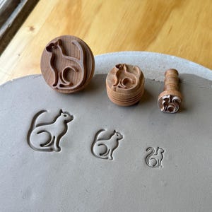 May include: Three round wooden stamps with cat-shaped relief designs are positioned above three corresponding cat-shaped impressions pressed into a flat, smooth slab of gray clay. The stamps vary in size, each featuring a stylized feline silhouette.
