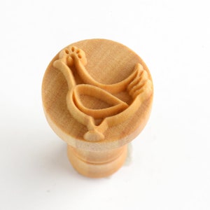 May include: A wooden cookie stamp featuring a raised design of a chicken. The stamp is light brown and has a round top with a cylindrical base.