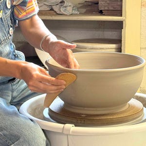 May include: A large, light gray ceramic bowl centered on a pottery wheel. A wooden rib tool is pressed against the outer rim of the bowl to smooth the clay. The bowl sits on a circular bat, and the entire assembly is contained within a white basin.
